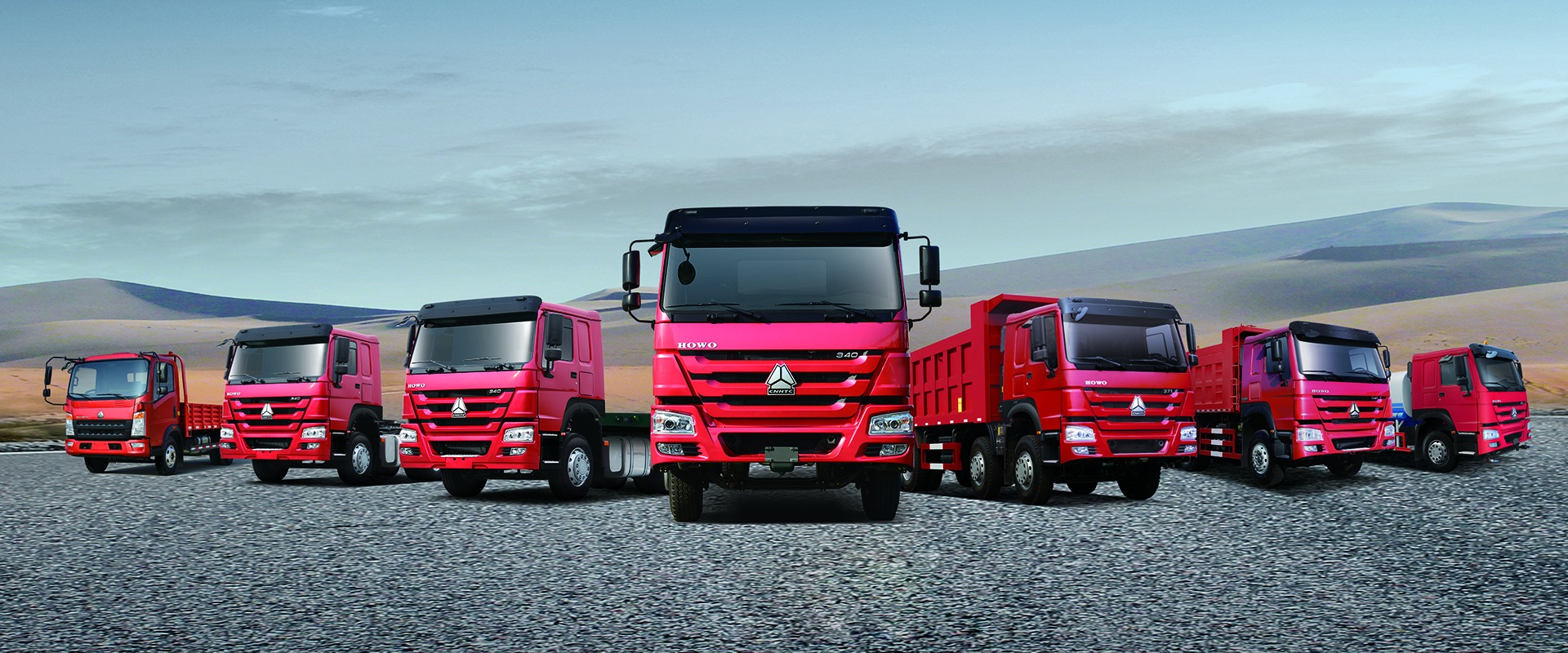 Multiple heavy trucks lined up at port ready for shipping, featuring Chinese heavy truck brands like Sinotruk or Shaanxi, blue sky background